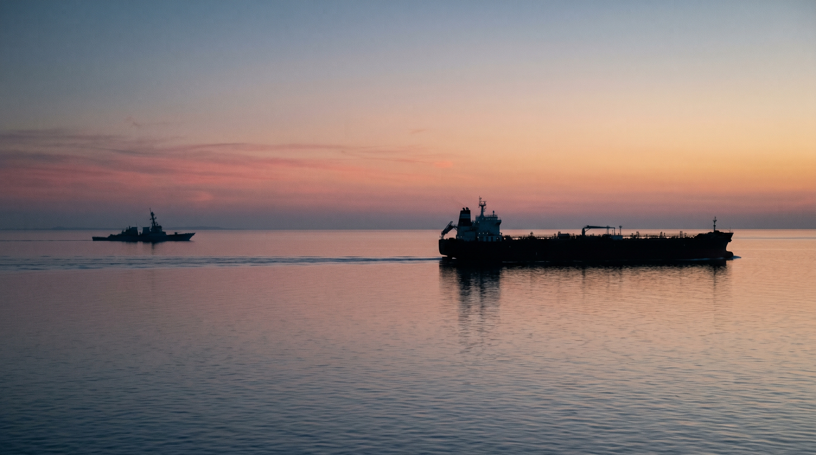 A wide telephoto shot of the Strait of Hormuz at first light, with a tanker silhouette crossing a narrow channel and a distant US naval vessel holding position on the Persian Gulf side