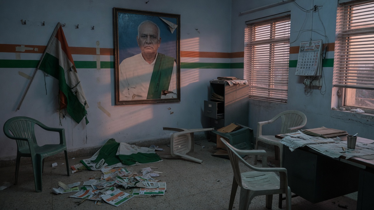 An empty political party office with a single large framed portrait of a leader on the wall, chairs in disarray as if recently vacated in argument, a torn banner on the floor